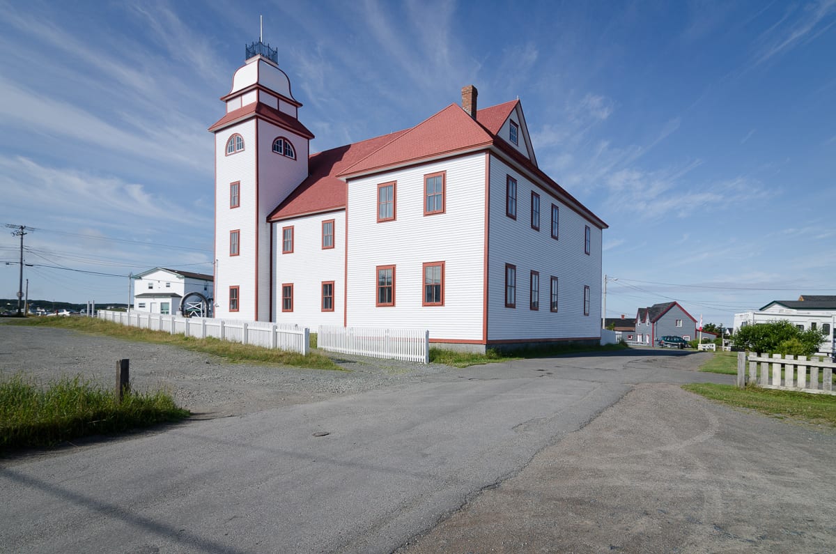 A large white clapboard hall with red trim, a hipped red roof and a tall domed corner tower, standing beside a gravel road at Bonavista, Newfoundland, under a blue sky
