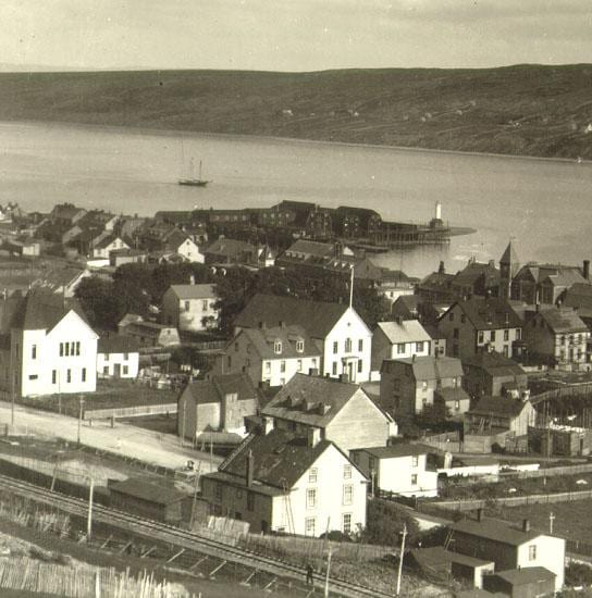 Sepia photograph of Harbour Grace, Newfoundland, around 1911, looking over wooden houses and a church spire toward a wharf and the harbour, with a railway line in the foreground