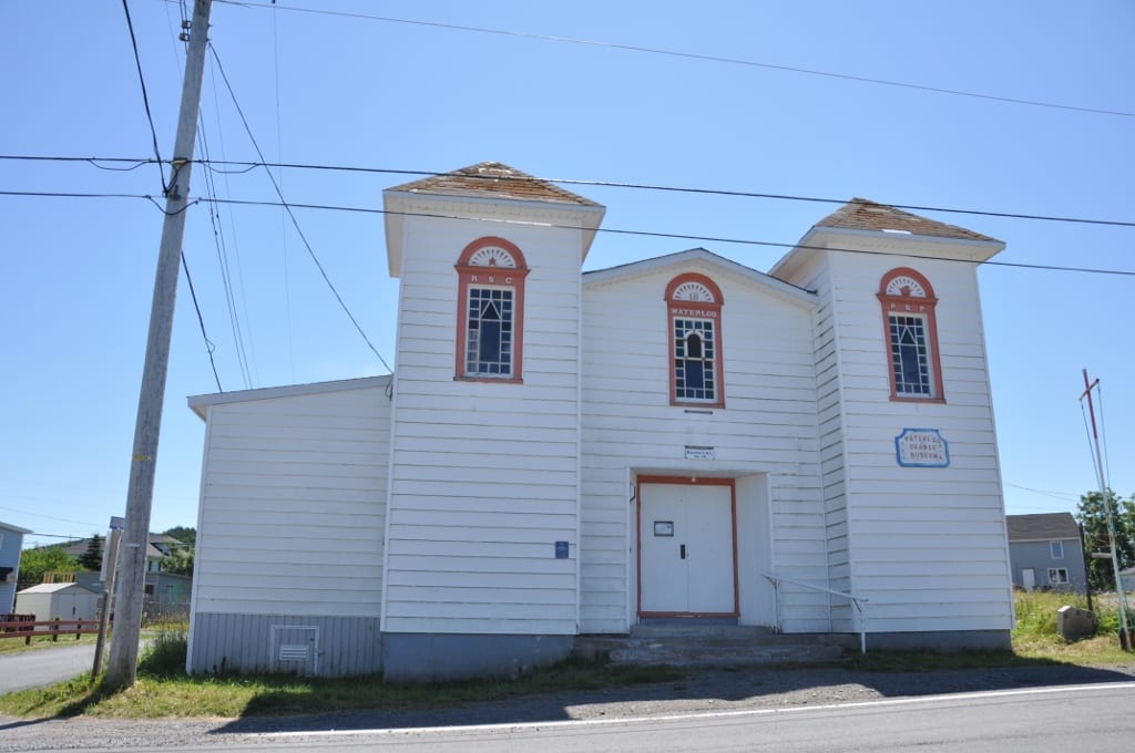 A white wooden lodge hall with two square towers, arched red-trimmed windows and a sign reading Waterloo Orange Museum, seen from the road at New Perlican, Newfoundland