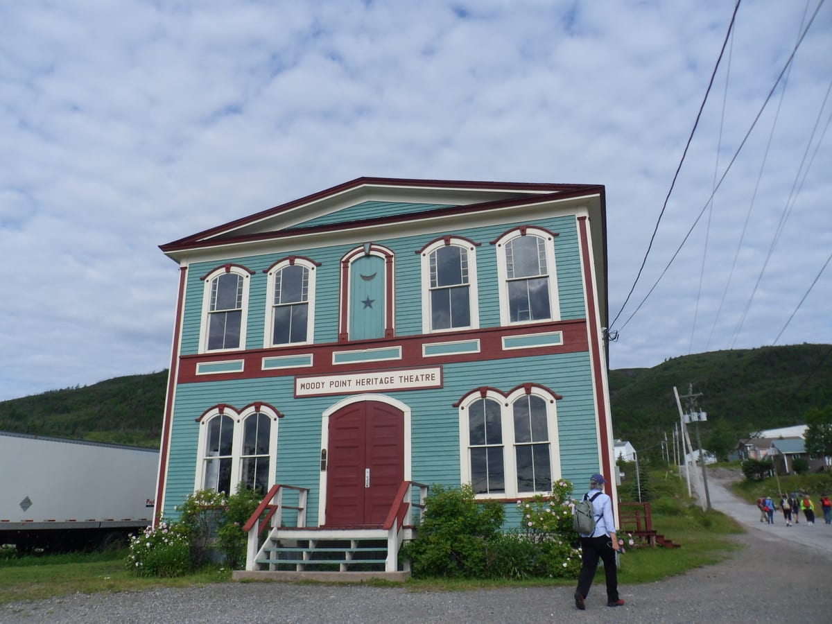 Two-storey teal clapboard hall with red trim and arched windows at Woody Point, Newfoundland, with a crescent and star above the upper central window and a sign reading Woody Point Heritage Theatre