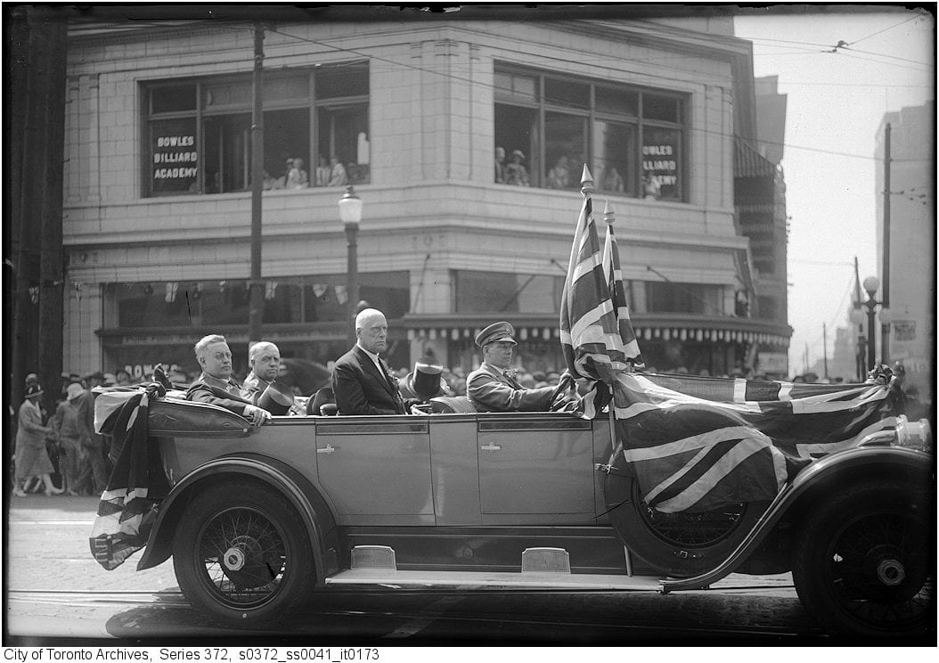 Four men in an open touring car draped with Union flags passing a corner building on a Toronto street, photographed on 12 July 1927
