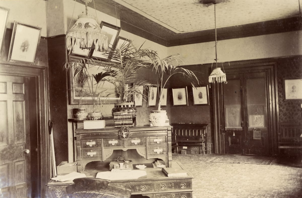 Interior of the mayor's office in Toronto's old city hall in 1898, with a large roll-top style desk, potted palms, hanging lamps and framed portraits along the walls